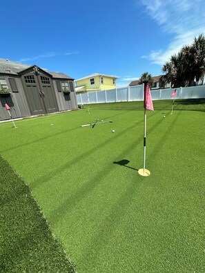 Um gramado artificial bem cuidado, com putting green e haste de sinalização, cercado por um muro branco e edifícios residenciais.