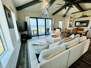 A spacious living room with a large sofa, a flat-screen TV, and a balcony with a view of the ocean. - Beautiful Oceanfront House with Pool in Flagler Beach (Flagler Beach)