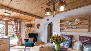 A rustic interior with a wooden ceiling, a stone wall, a green armchair, a wooden dining table with a placemat, and a vase of flowers. - Chalet Hüttenzauber (Kirchberg in Tirol)