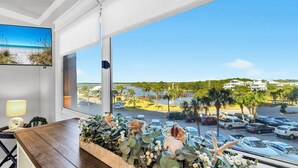 A room with a large window overlooking a beach and parking area, a wooden table with a plant arrangement, and a lamp on the side. - Beachfront Gem Lakeside Resort Amenities Near 30A (Panama City Beach)