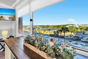Ett rum med ett stort fönster med utsikt över en strand och en parkeringsplats, ett träbord med en blomsteruppsättning och en lampa i hörnet.