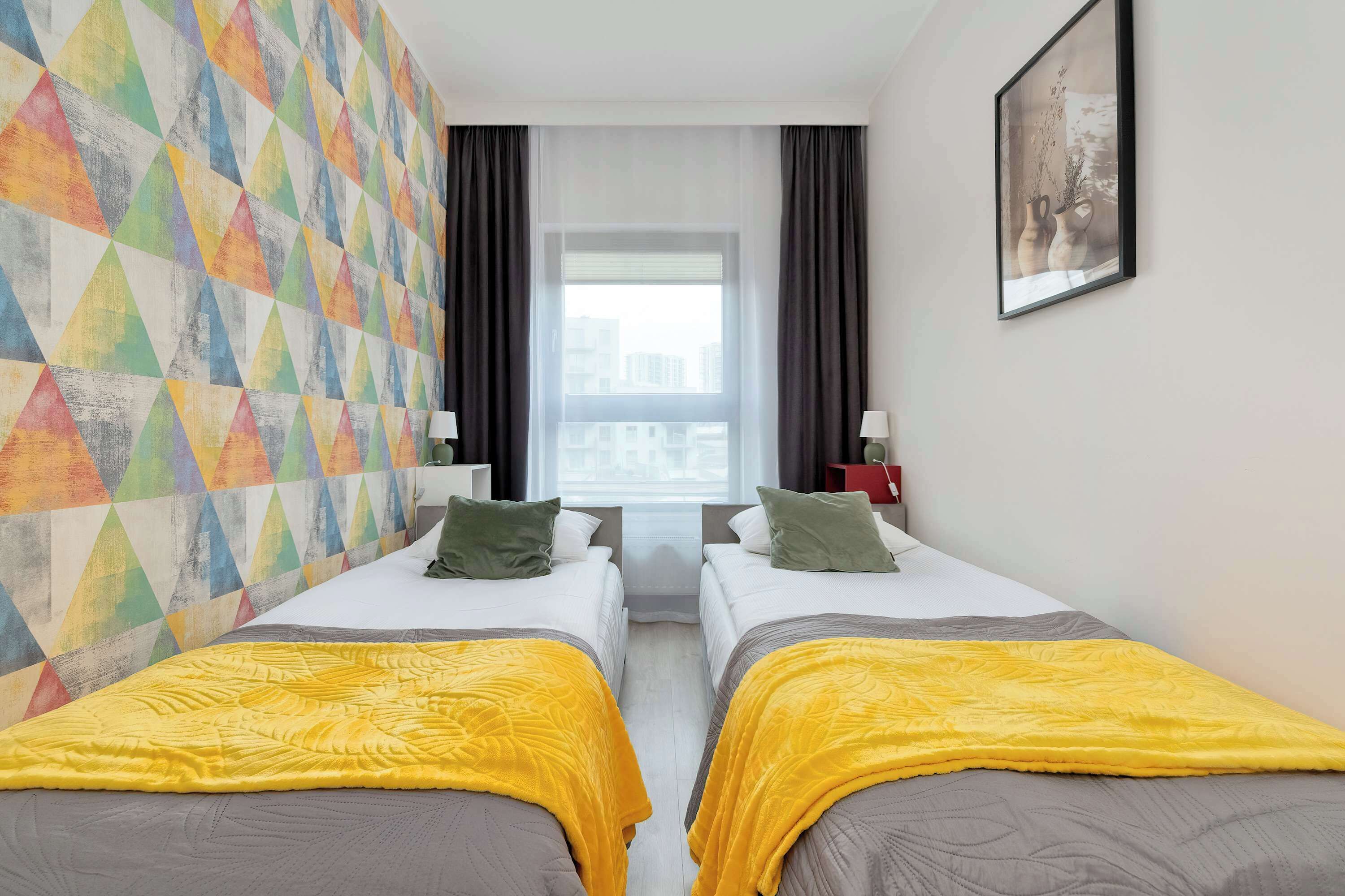 Habitación de hotel con dos camas, un muro con diseño geométrico y colores vivos, un ventanal grande y un cuadro en la pared.
