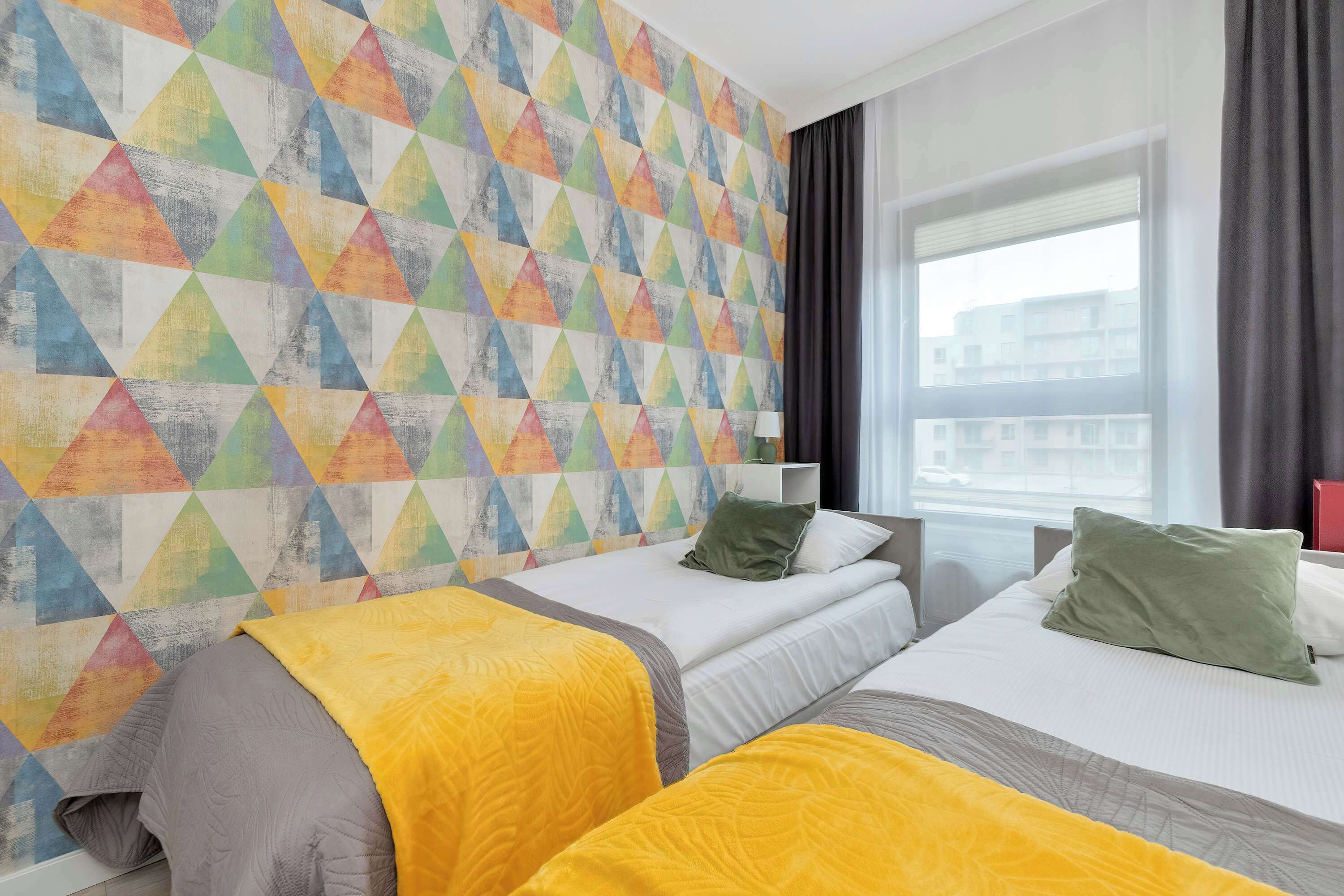 A hotel room with two beds, a colorful geometric wall, a window with curtains, and a view of buildings outside.