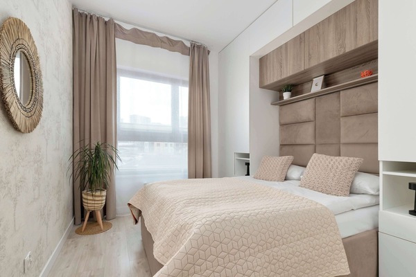 A double bedroom is shown with a large round mirror reflecting the light from the opposite window. The room features a built-in wooden headboard unit that matches the flooring and closet.
