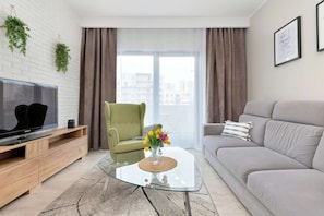 Ein modernes Wohnzimmer mit Fernseher, einem grünen Sessel, einem grauen Sofa und einem Glastisch mit Blumenvase.