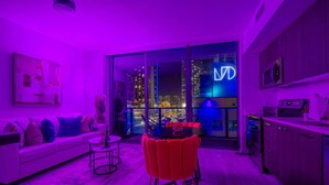 A modern living room with a sofa, a dining table with red chairs, and a kitchen area with a microwave. A large window offers a view of the city at night. - Brickell Area| Free Parking| Balcony| Rooftop Pool (Miami)