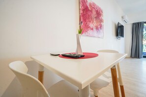 Ein weißer Tisch mit Fernbedienung und Pflanze auf einer roten Matte, weiße Stühle, ein pinkes abstraktes Gemälde, ein Fernseher und ein Fenster mit Vorhängen.
