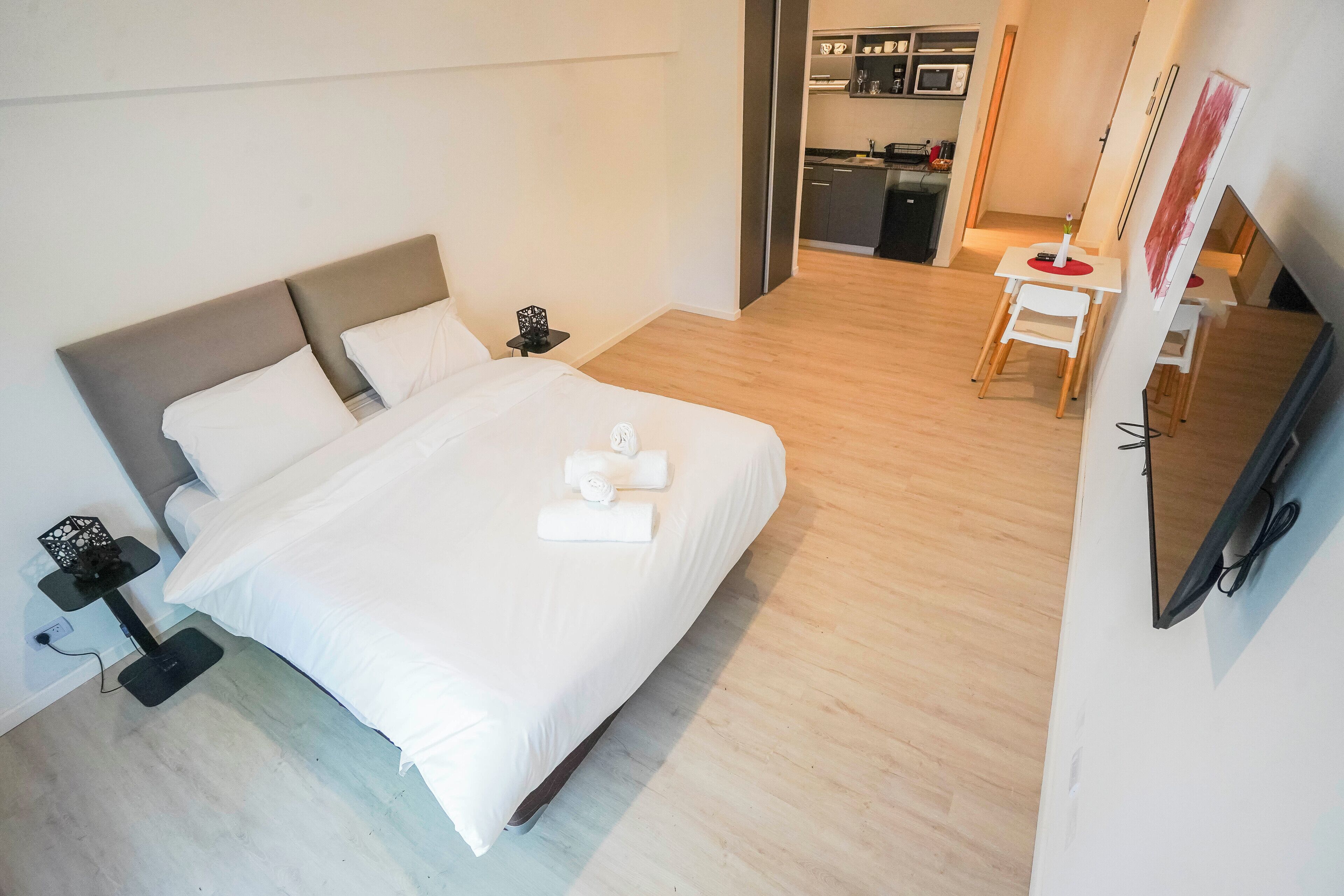 A modern hotel room with a large bed, a desk, a chair, a TV, and a kitchen area in the background.