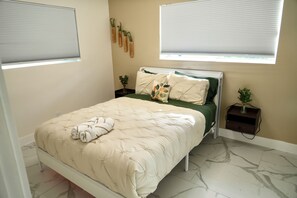 A bedroom with a bed, a nightstand, a window with blinds, and a wall-mounted shelf with plants. - Hollywood 1811 (Hollywood)