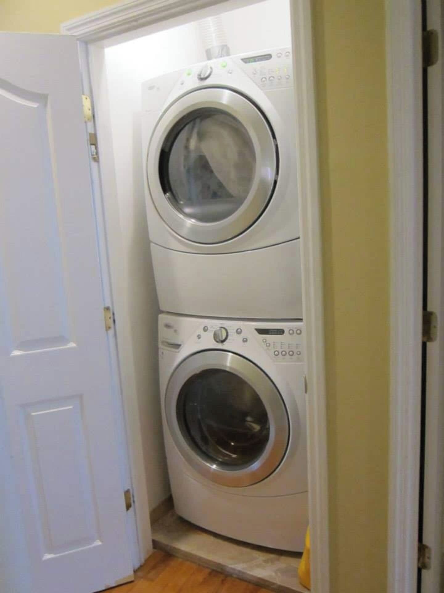 Un appareil compact combinant une machine à laver et un sèche-linge à chargement frontal, doté de deux cuves, installé dans un petit espace buanderie avec une porte blanche et un sol carrelé.