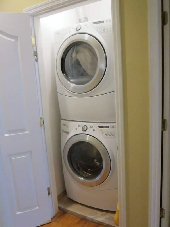 A compact, two-basin front-loading washer and dryer unit in a small laundry room with a white door and tiled floor.