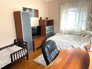 A bedroom with a bed, a desk with a computer, a chair, a wardrobe, and a window with curtains.