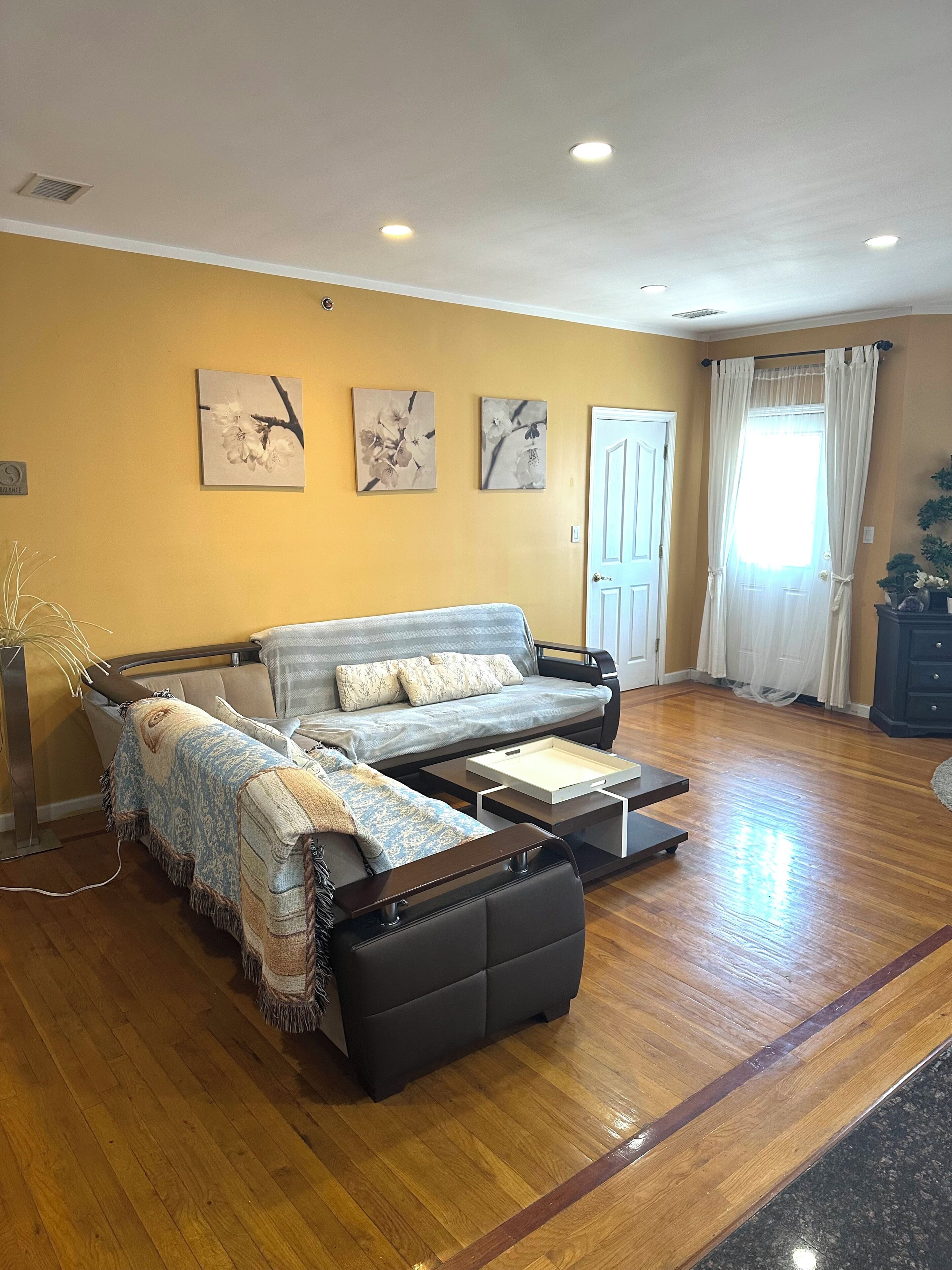 A living room with a sofa, a coffee table, and a side table. There are three wall-mounted pictures, a door, and a window with curtains.
