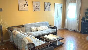 A living room with a sofa, a coffee table, and a side table. There are three wall-mounted pictures, a door, and a window with curtains.