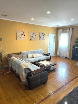 Una sala de estar con sofá, una mesa de centro y una mesita auxiliar. Hay tres cuadros colgados en la pared, una puerta y una ventana con cortinas.