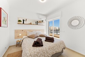 Une chambre à coucher comprenant un lit, une table de chevet, une étagère murale avec des plantes, un miroir rond et une fenêtre donnant sur des immeubles.