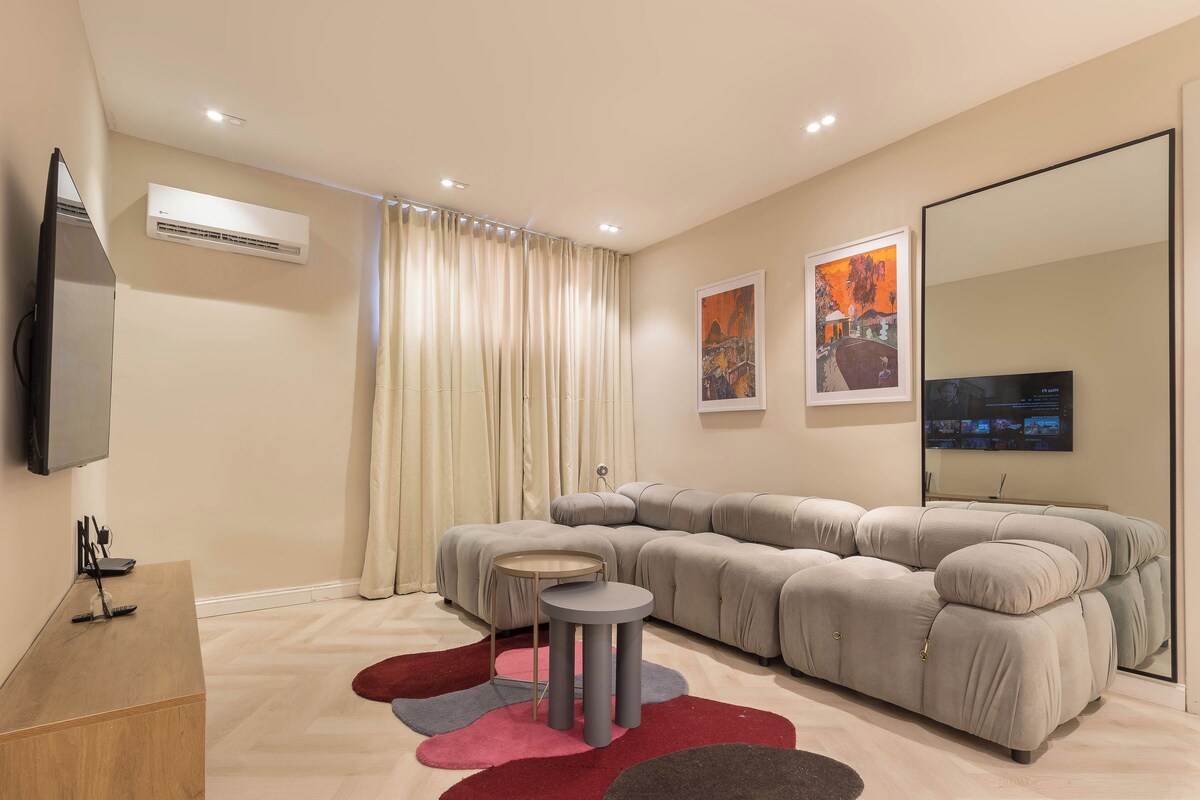 A modern living room with a sectional sofa, a small round table, a flat-screen TV, and a large mirror.
