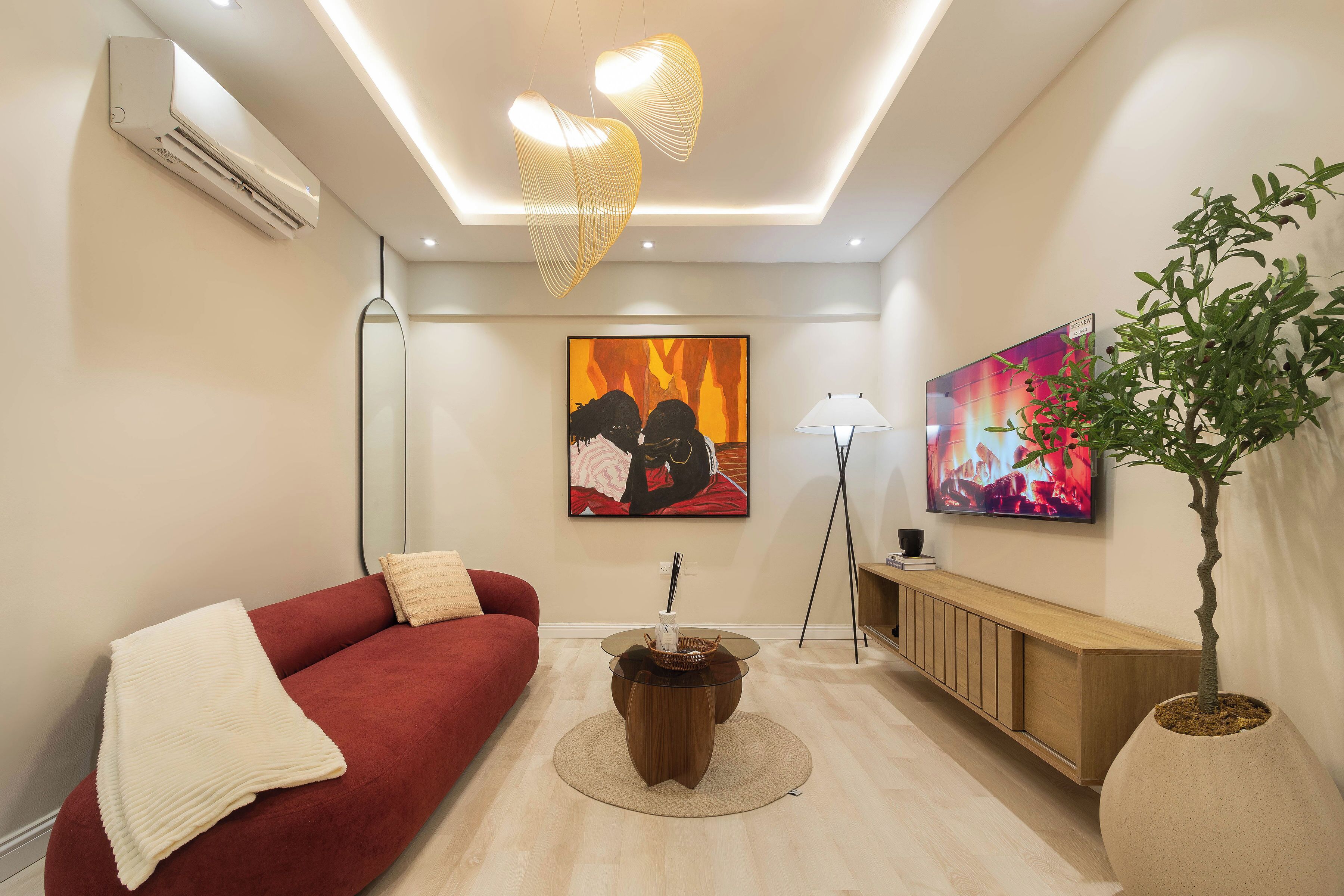 Ein modernes Wohnzimmer mit einem roten Sofa, einem runden Holztisch, einem großen Gemälde, einer Topfpflanze und einem an der Wand befestigten Fernseher.
