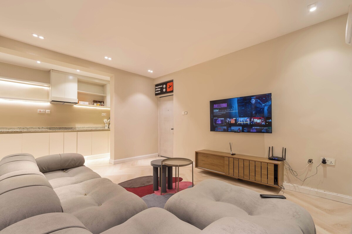 A modern living room with a flat-screen TV, a sofa, a small round table, and a kitchen area with cabinets and a countertop.