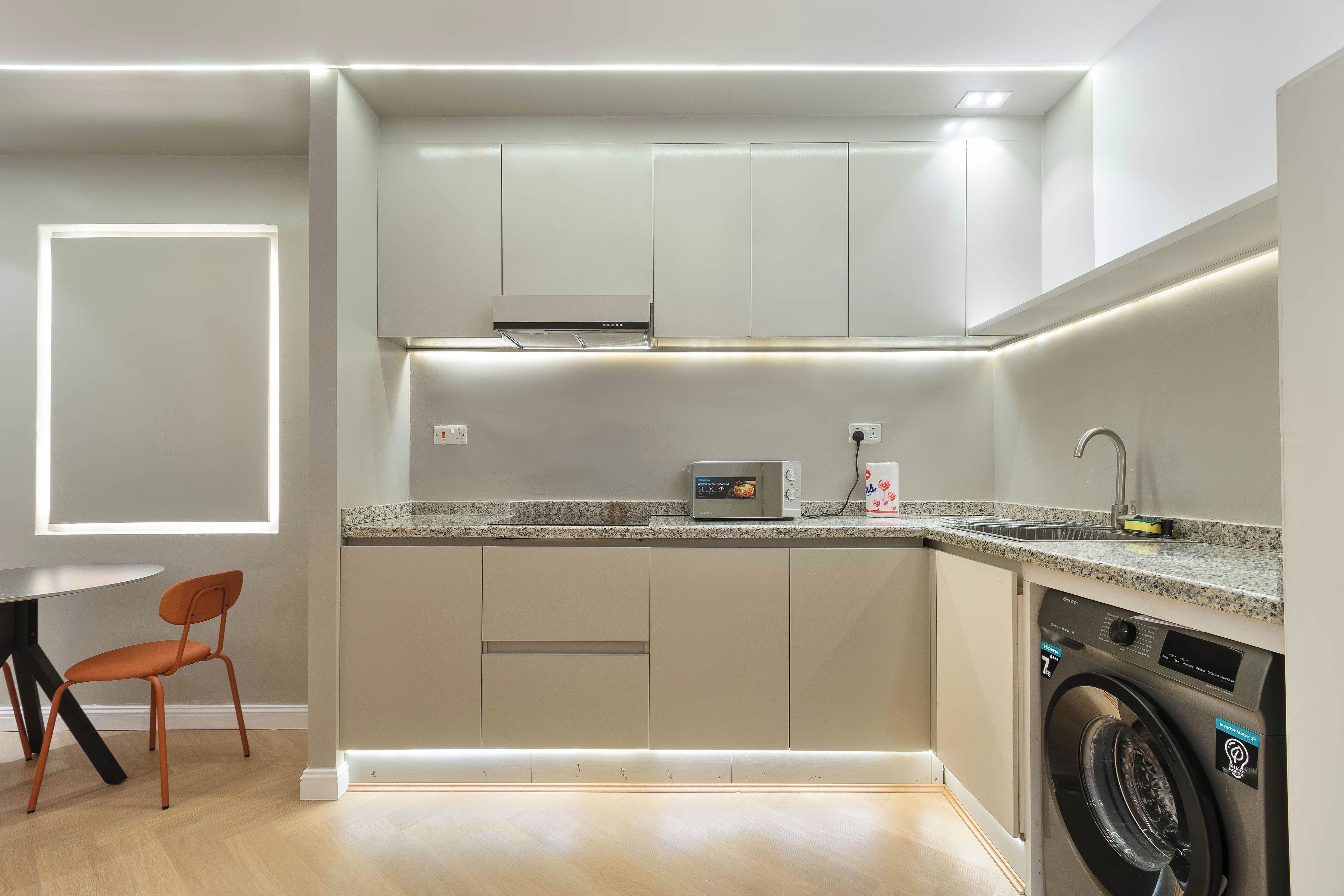 A compact kitchen with a microwave, washing machine, and a small dining table with a chair.
