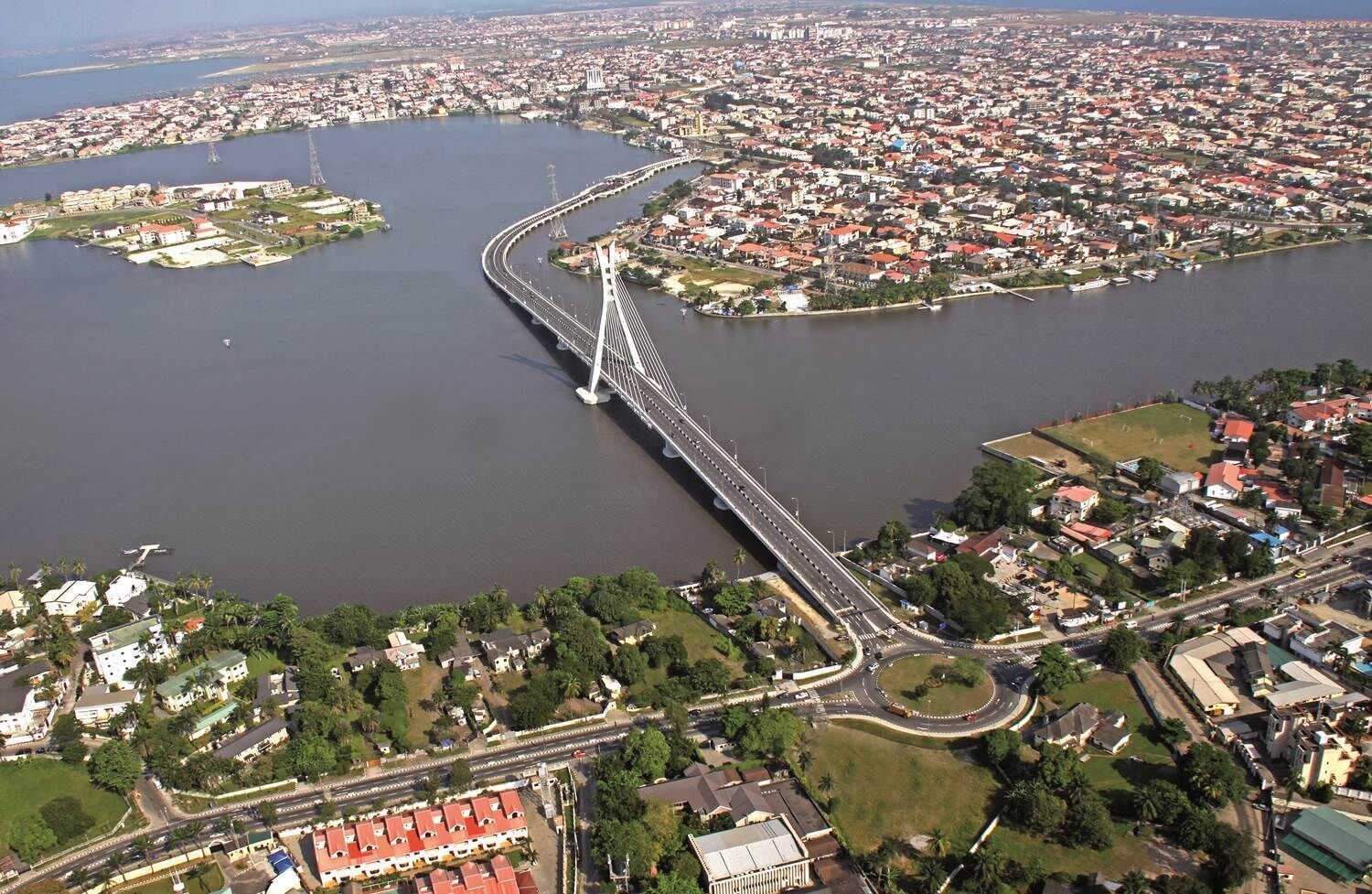 An aerial view of a city with a prominent bridge crossing a river, surrounded by residential areas and greenery.