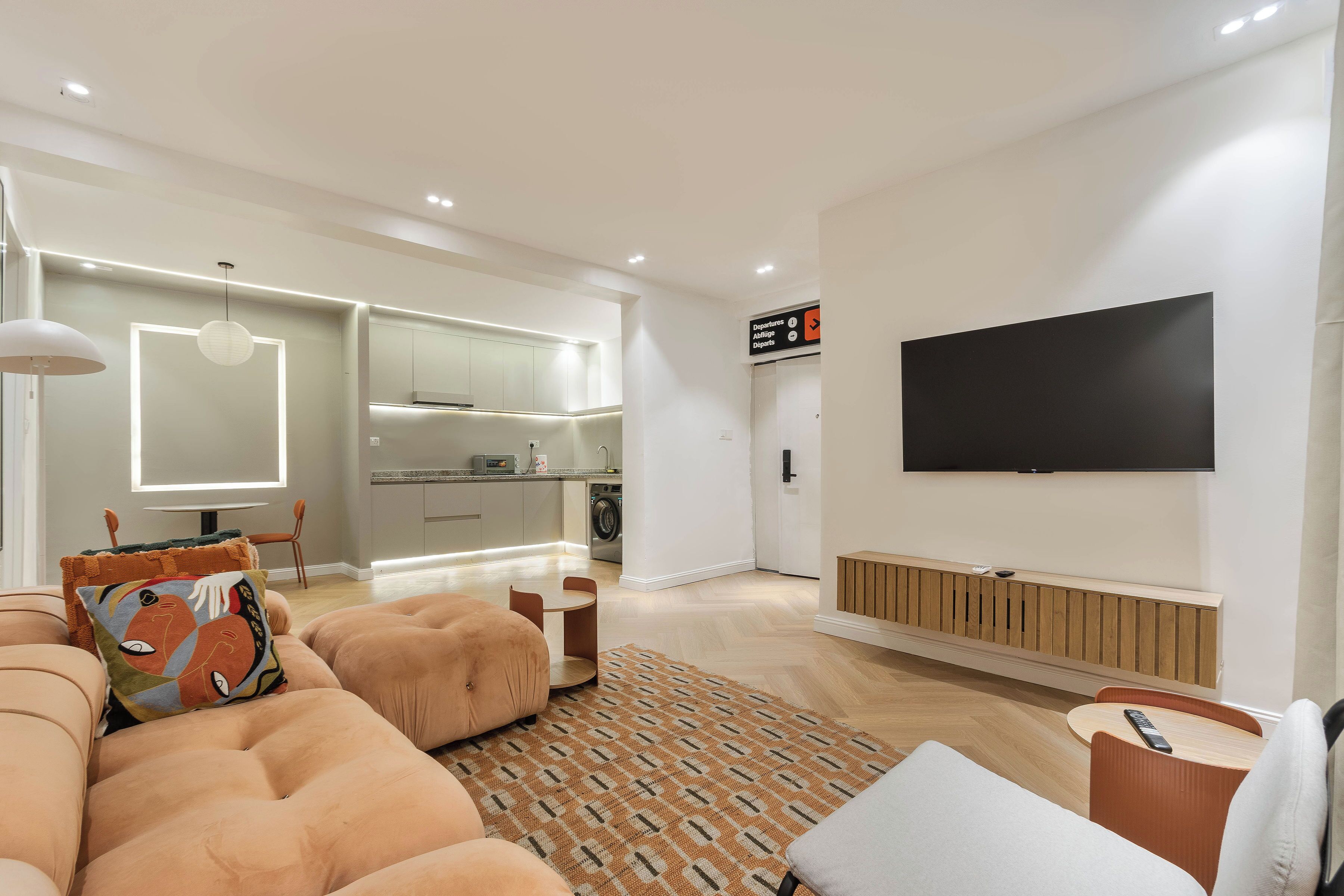 A modern living room with a sofa, ottoman, and a patterned rug. A flat-screen TV is mounted on the wall, and a kitchen is visible in the background.