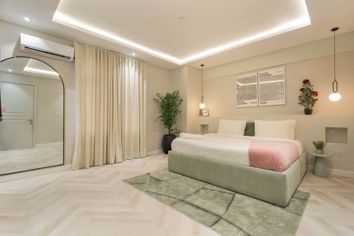 A modern bedroom with a large bed, a mirror, a potted plant, and a wall-mounted artwork.