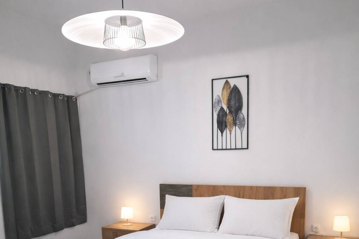 Un dormitorio moderno con una cama grande, cabecera de madera, dos mesitas de noche con lámparas, aire acondicionado empotrado en la pared y una obra de arte con una hoja enmarcada.