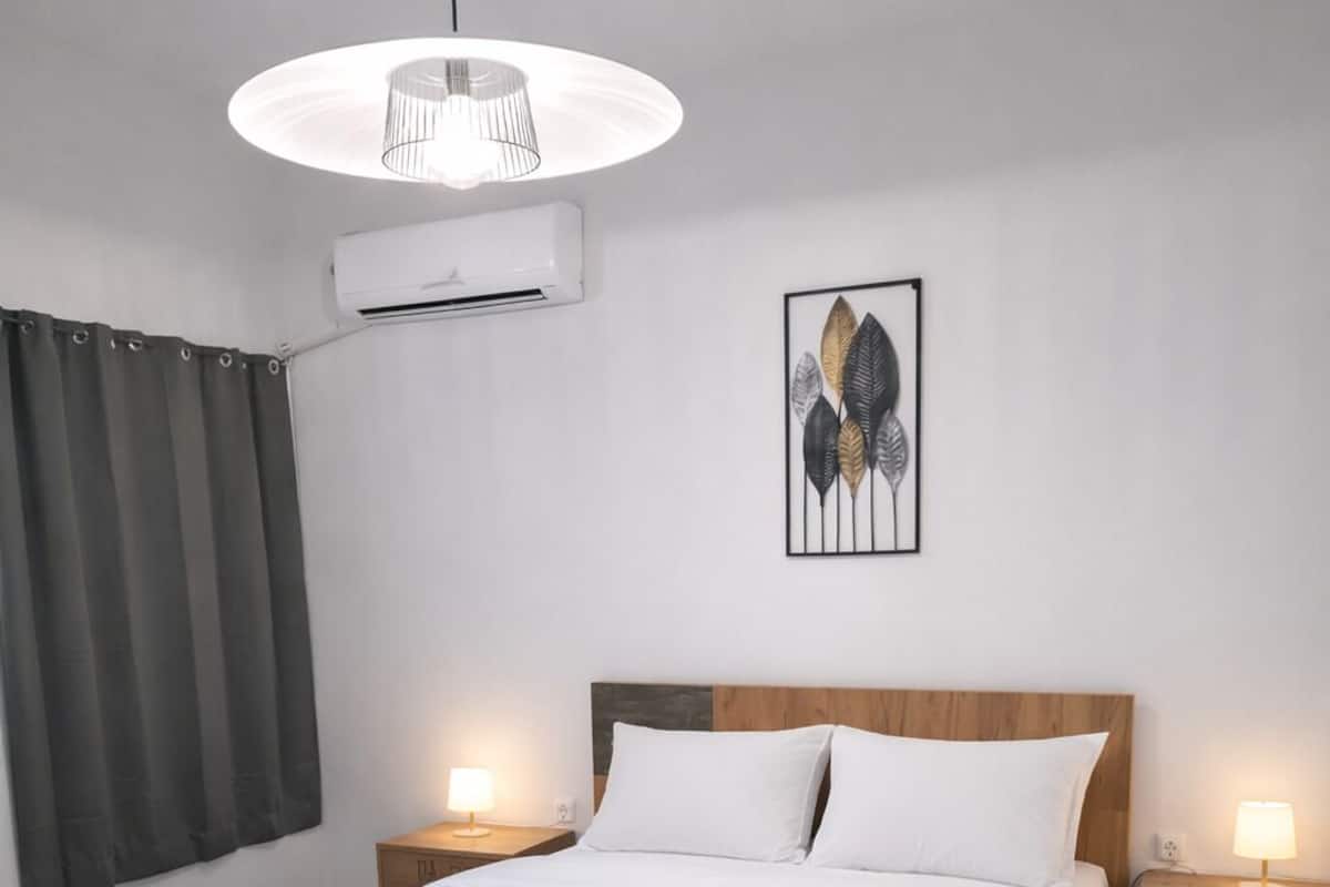 Un dormitorio moderno con una cama grande, cabecera de madera, dos mesitas de noche con lámparas, aire acondicionado empotrado en la pared y una obra de arte con una hoja enmarcada.