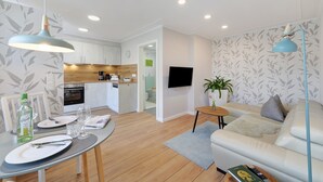 A modern living room with a sofa, a dining table set for two, a kitchen in the background, and a flat-screen TV mounted on the wall. - Apartment 'Ferienwohnungen Meerblick -' with Private Terrace, Garden View, and Wi-Fi (Bastorf)