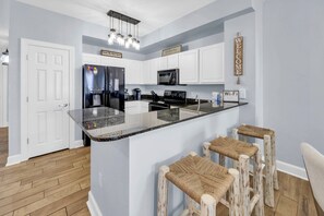 A kitchen with white cabinets, a black refrigerator, a microwave, and a coffee maker. There is a bar area with stools and a 'Welcome' sign. - ☀️Stunning Gulf Views | 2 Pools + 2 Hot Tubs | Sunrise Beach #1010☀️ (Panama City Beach)