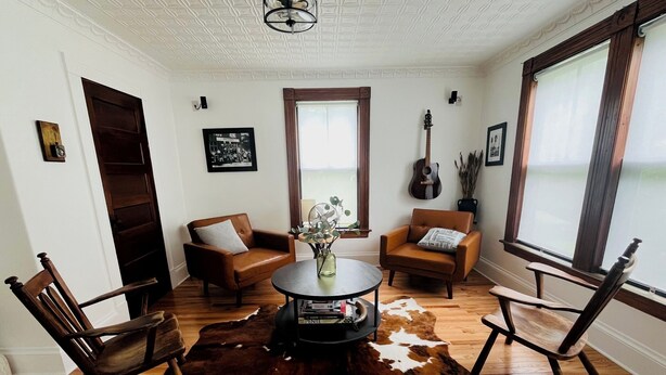 Une pièce avec un plancher en bois, deux fauteuils en cuir, une table basse ronde, un tapis en peau de vache, une guitare et une fenêtre avec des rideaux.