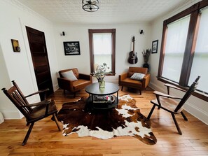 A room with a wooden floor, two leather armchairs, a round coffee table, a cowhide rug, a guitar, and a window with curtains. - Lakeport Sound House (Laconia)