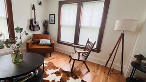 Une pièce avec un plancher en bois, un canapé marron, une table recouverte d’un tapis en peau de vache, une guitare, une lampe et une fenêtre avec des stores.