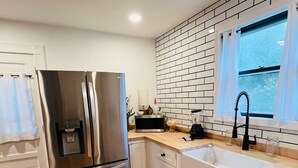 Une cuisine moderne dotée d’un réfrigérateur en acier inoxydable, de meubles blancs, d’un pare-feu en briques et d’une fenêtre avec des rideaux.