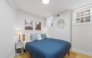 Un dormitorio con cama, mesita de noche, lámpara, ventana con persianas y espejo montado en la pared.