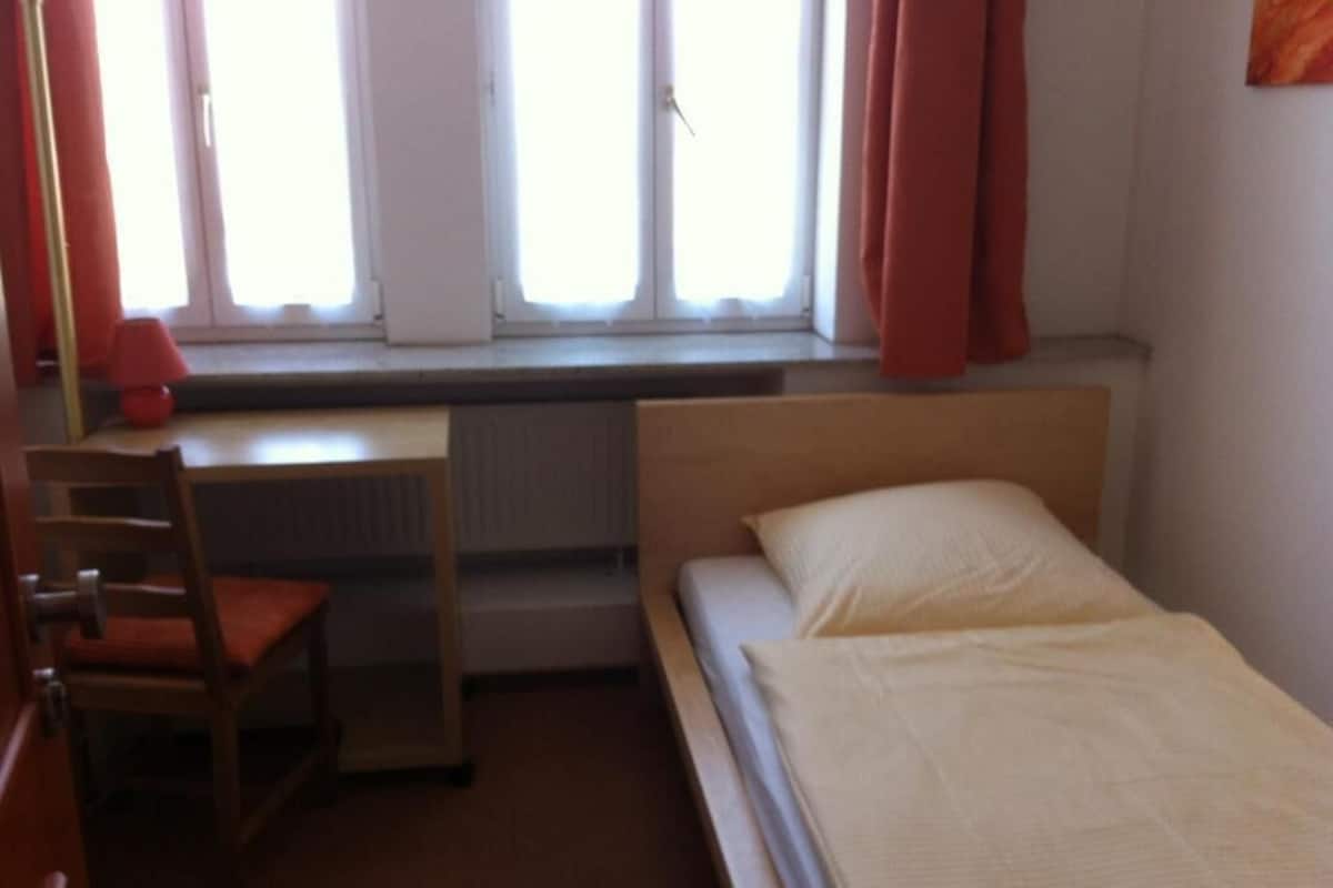 A single bed with a wooden headboard, a desk with a chair, and a radiator under a window with curtains.