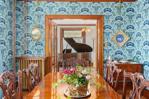 Una sala da pranzo con un pianoforte, sedie di legno, un tavolo con un centrotavola floreale e carta da parati decorata.