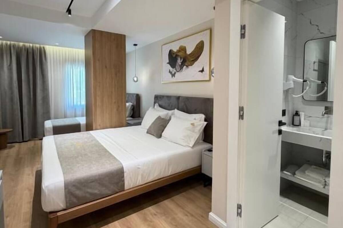 Un dormitorio moderno con una cama grande, piso de madera, un cuadro en la pared y un baño con espejo.