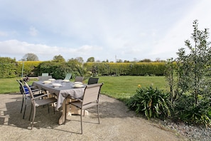 Un espace extérieur aménagé pour les repas, avec une table et des chaises, entouré de verdure et d’une haie.