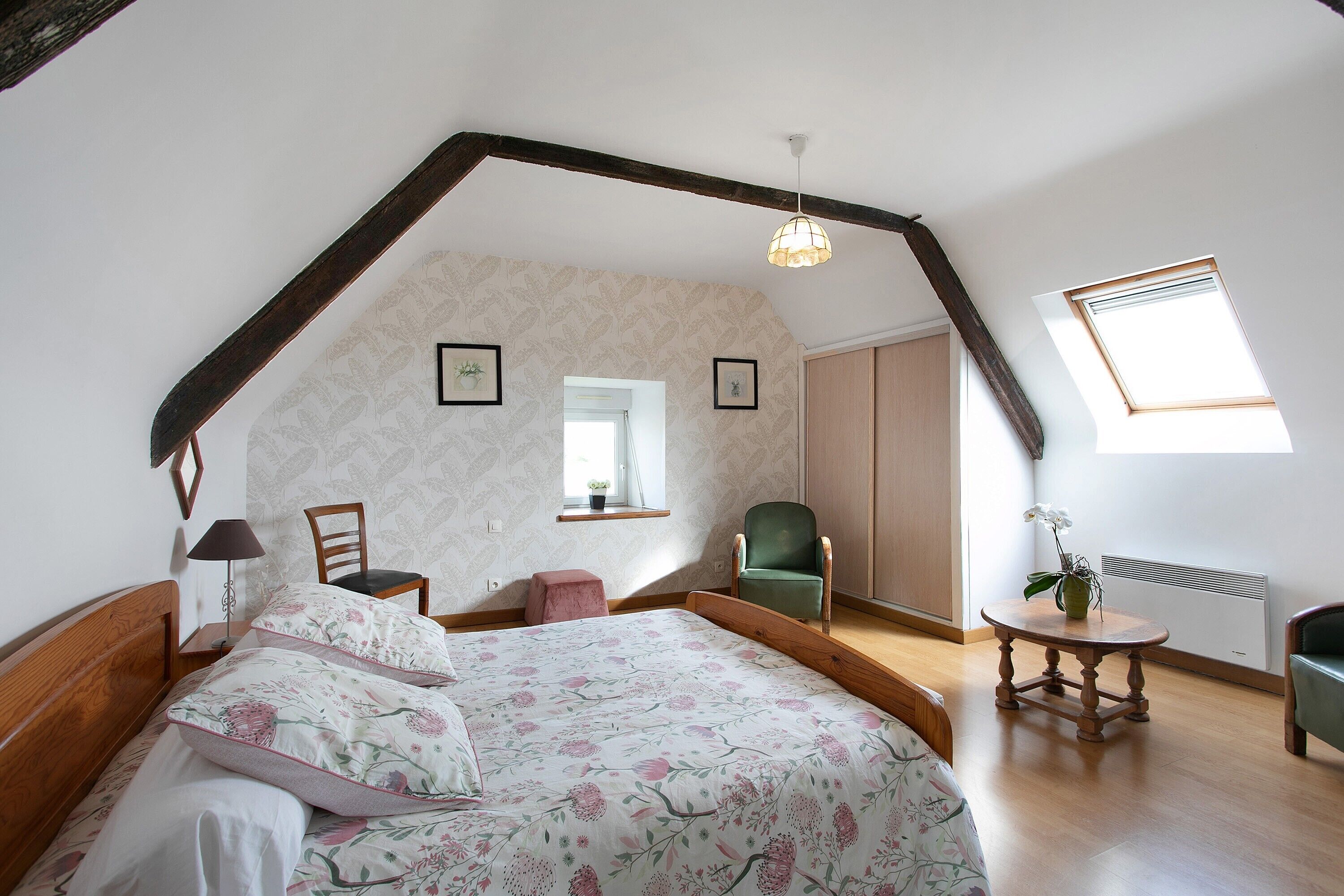 Une chambre avec un plafond en pente, un lit avec une literie à motifs floraux, une chaise, une petite table avec une plante et une fenêtre donnant sur un beau paysage.