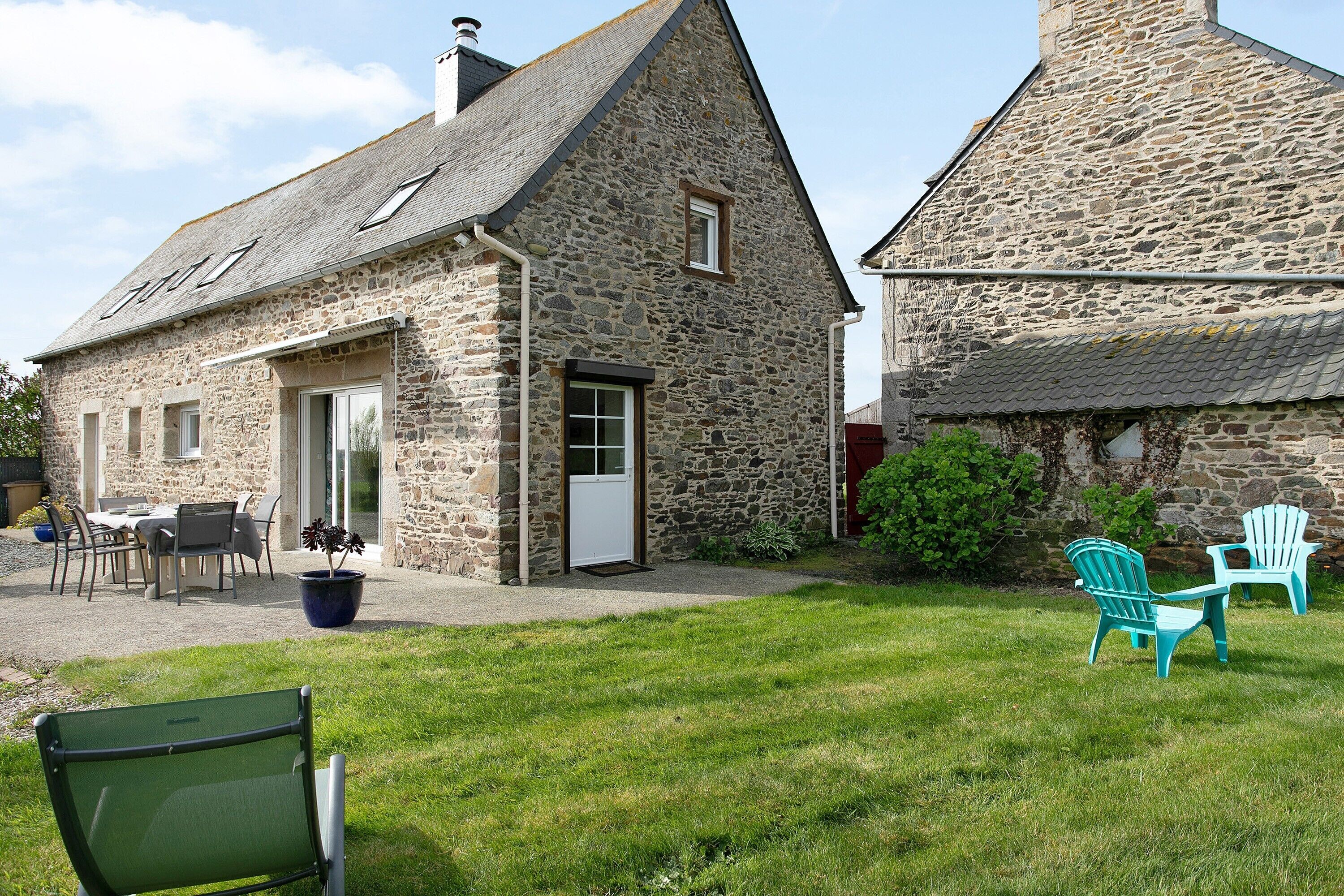 Un bâtiment en pierre doté d’une porte, de fenêtres et d’un petit espace jardin aménagé avec des chaises et un banc.