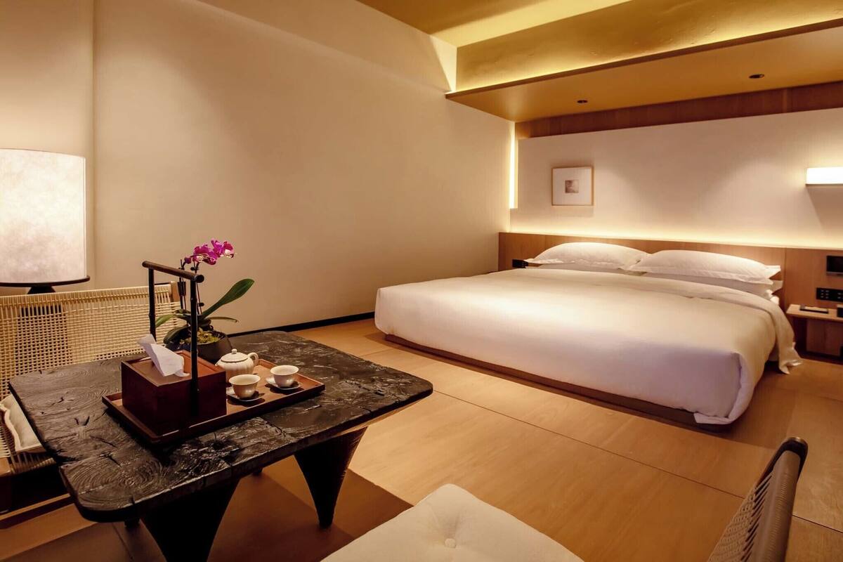 A hotel room with a bed, bedside table with a tray of tea, a potted orchid, and a wall-mounted TV.