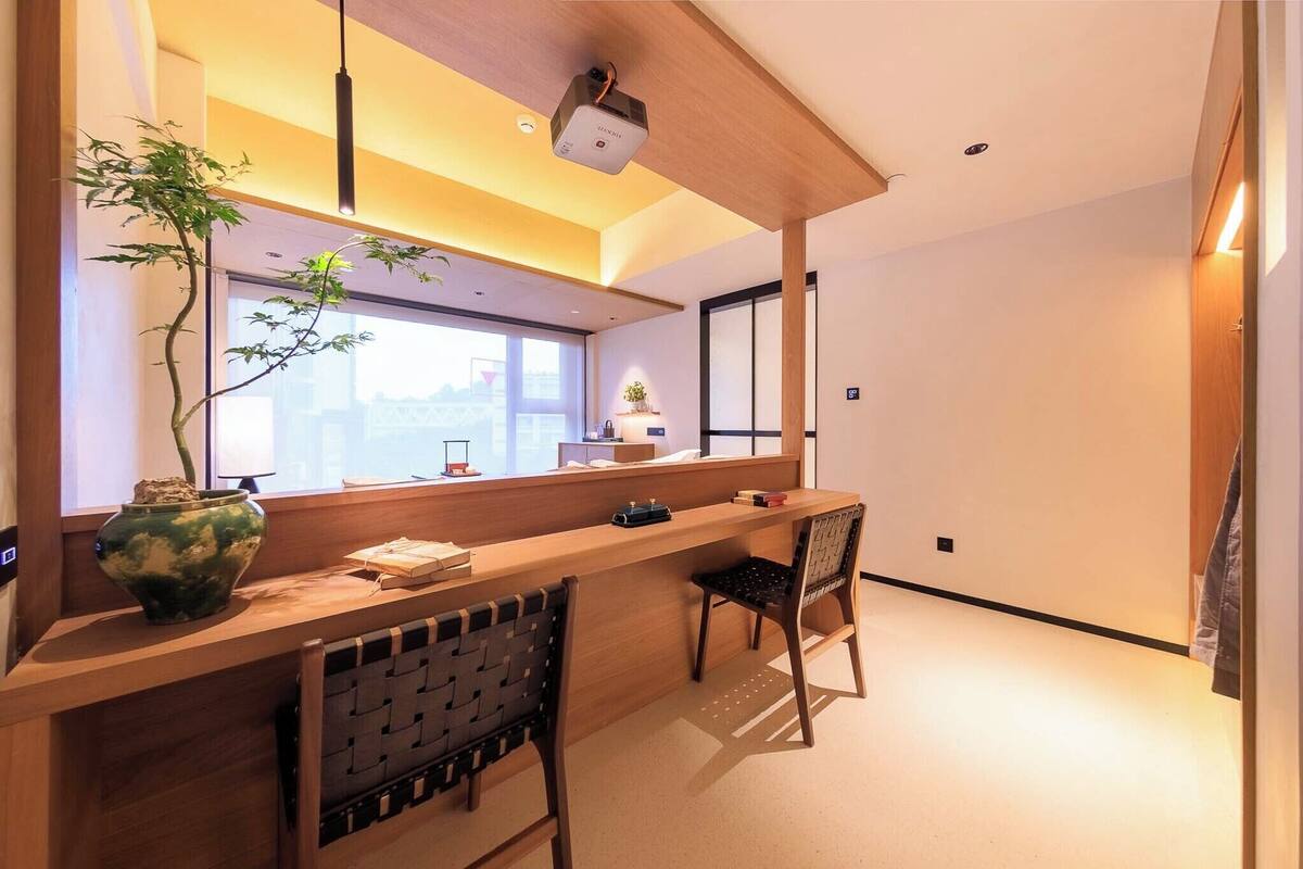 A modern hotel room with a wooden reception desk, a potted plant, a chair, and a large window.