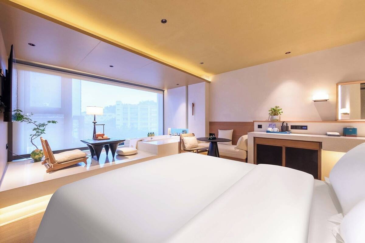 A modern hotel room with a large bed, a desk, a chair, a sofa, and a large window offering a city view.