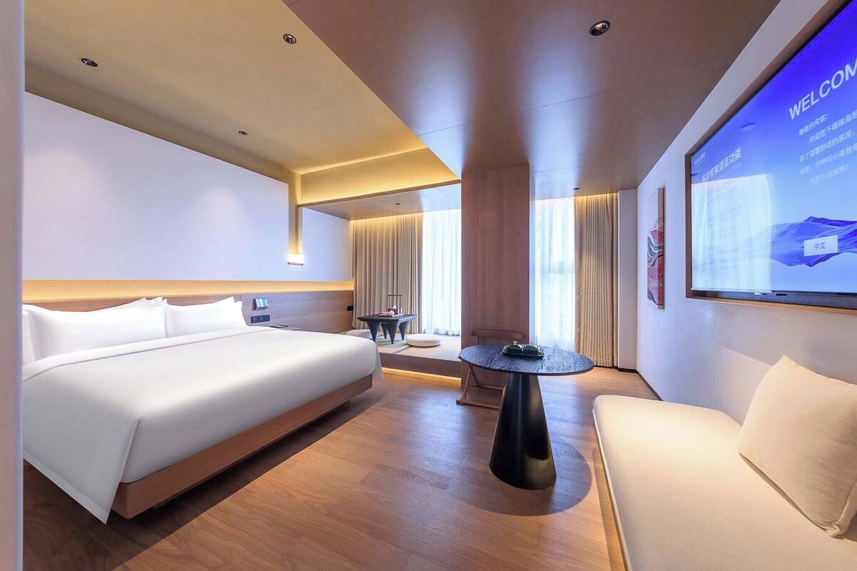 A modern hotel room with a large bed, a sofa, a small table, and a wall-mounted screen displaying 'WELCOME'.