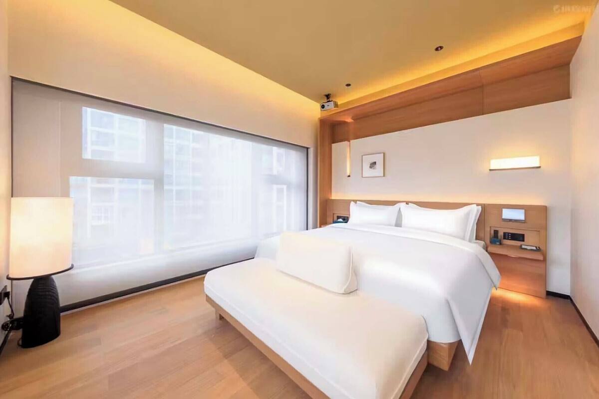A hotel room with a large bed, a bedside table, a lamp, a nightstand, and a view of buildings through the window.