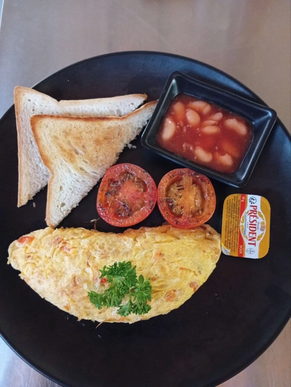 Um prato com torradas, feijão, tomate, um pequeno recipiente de manteiga e uma omelete.