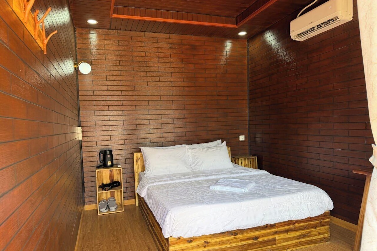 A wooden-framed bed with white bedding, a small wooden shelf with a coffee maker and shoes, and a wall-mounted air conditioner.