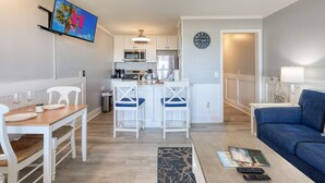 Een moderne keuken met witte kasten, een magnetron en een koffieapparaat. Er is een eethoek met een houten tafel en witte stoelen. In de woonkamer staat een blauwe bank en een klein vloerkleed.